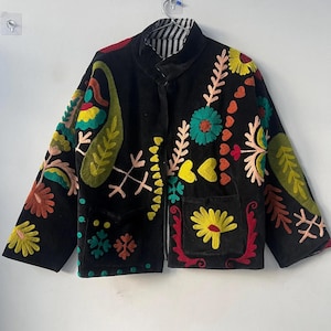 Puede incluir: Chaqueta negra con bordados florales coloridos. La chaqueta tiene cuello alto, dos bolsillos delanteros y forro a rayas. El bordado incluye flores, hojas y corazones en tonos rojos, amarillos, verdes y azules.