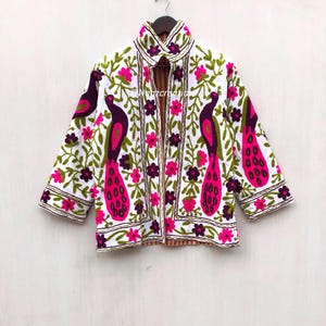 May include: White jacket with a vibrant peacock and floral embroidery design. The jacket features a collared neckline and three-quarter sleeves. The embroidery includes pink, purple, green, and white colors. The jacket is hanging on a wooden hanger.