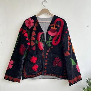 Peut inclure: Veste en velours noir avec broderies florales colorées rouges, roses, vertes et oranges. La veste a deux poches avant et une doublure rayée. La veste est suspendue à un cintre en bois.