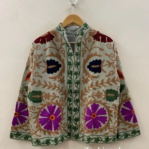 Puede incluir: Chaqueta de color claro con intrincados bordados florales en tonos morado, marrón, verde y azul marino. La chaqueta tiene un cuello alto y una parte delantera abotonada. El texto "suzanifashionshop" es visible en la parte inferior.