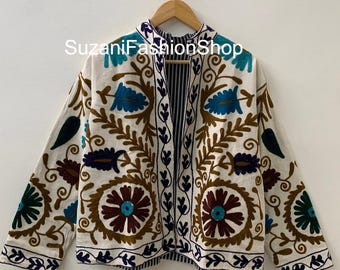 Puede incluir: Chaqueta bordada de color crema con motivos florales en tonos marrón, azul y verde azulado. La chaqueta presenta mangas largas y un forro a rayas. El texto "SuzaniFashionShop" es visible en la chaqueta.