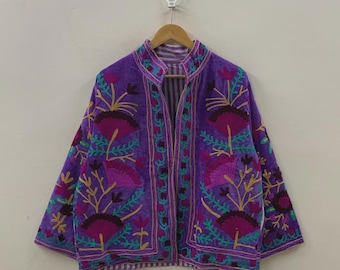 May include: A purple embroidered jacket with a mandarin collar. The jacket features a floral design in shades of pink, green, and gold. The jacket is hanging on a wooden hanger against a plain white background.