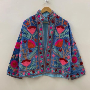 May include: A blue embroidered jacket with a high collar and long sleeves. The jacket features a floral design in shades of pink, orange, and green. The jacket is hanging on a wooden hanger against a white wall.