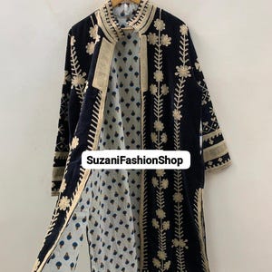 May include: A long, open-front black coat with cream-colored floral embroidery. The coat has a patterned interior with blue and cream designs and the text "SuzaniFashionShop". It is suspended from a wooden hanger.