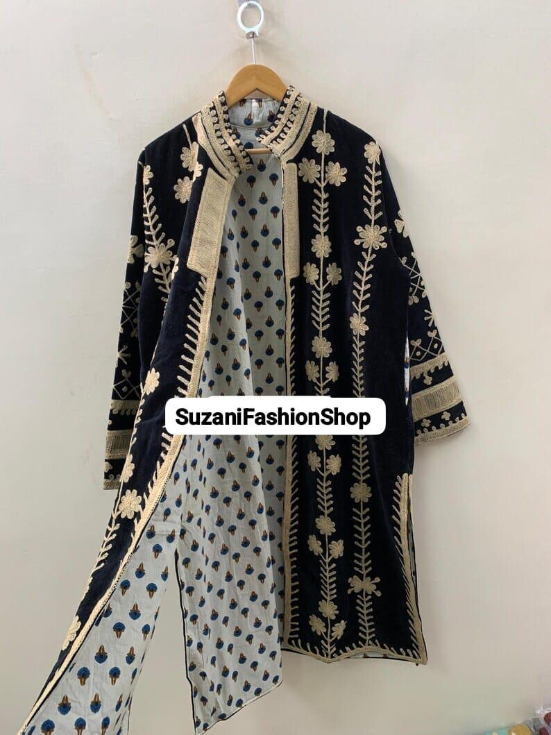 May include: A long, open-front black coat with cream-colored floral embroidery. The coat has a patterned interior with blue and cream designs and the text "SuzaniFashionShop". It is suspended from a wooden hanger.