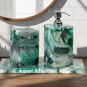 Puede incluir: Un juego de baño que incluye un dispensador de jabón con bomba plateada, un vaso y una bandeja. El juego está hecho de piedra veteada verde y blanca. El texto "Asian Mason's Art" es visible en la bandeja.
