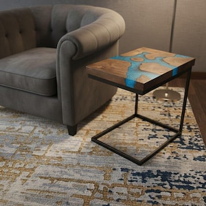 Puede incluir: Una mesa auxiliar moderna con una tapa de madera con un diseño de río de resina azul. La mesa tiene una estructura de metal negro y una forma rectangular. La mesa está al lado de un sillón gris.
