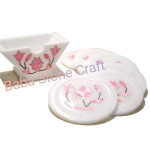 Può includere: Set di sottobicchieri in marmo bianco con un portabicchieri quadrato abbinato. Ogni sottobicchiere e il portabicchieri sono decorati con intarsi floreali rosa. Il testo "Baba Stone Craft" è visibile.