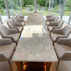 Peut inclure: Une grande table à manger rectangulaire avec une surface grise clair à motifs, entourée de douze chaises rembourrées grises claires. La table a des pieds en bois. La pièce a de grandes fenêtres donnant sur un jardin.