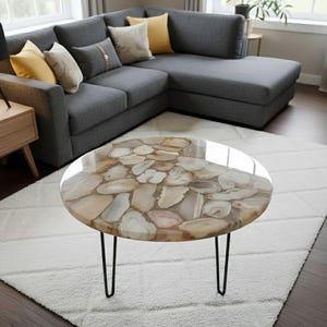 May include: A round coffee table with a polished agate stone top and black hairpin legs. The table sits on a white shag rug in front of a gray sectional sofa with yellow and beige throw pillows. The room has hardwood floors and a window.