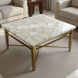 Puede incluir: Mesa de centro cuadrada con tapa blanca y dorada estampada y patas de metal dorado. La mesa tiene el texto "Asian Mason's Art" en la parte superior. La mesa está en un salón.