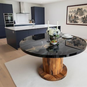 Puede incluir: Una mesa de comedor redonda y negra con una base de madera única. La superficie de la mesa tiene un aspecto brillante y veteado. La mesa está decorada con velas, flores y platos, lo que sugiere un ambiente de comedor. La cocina tiene armarios azul marino y una encimera blanca.