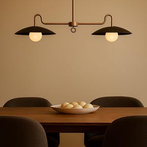 May include: A modern dining room scene featuring a dark wood table and four dark gray chairs. Above the table hangs a two-light pendant with black shades and spherical bulbs. A white bowl holds several round, golden-brown rolls.