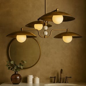 May include: A brass-toned chandelier with four lampshades and globe lights. The fixture has a central stem and curved arms. The lampshades are a flat, disc shape. The fixture is above a bathroom vanity with a mirror and sink.
