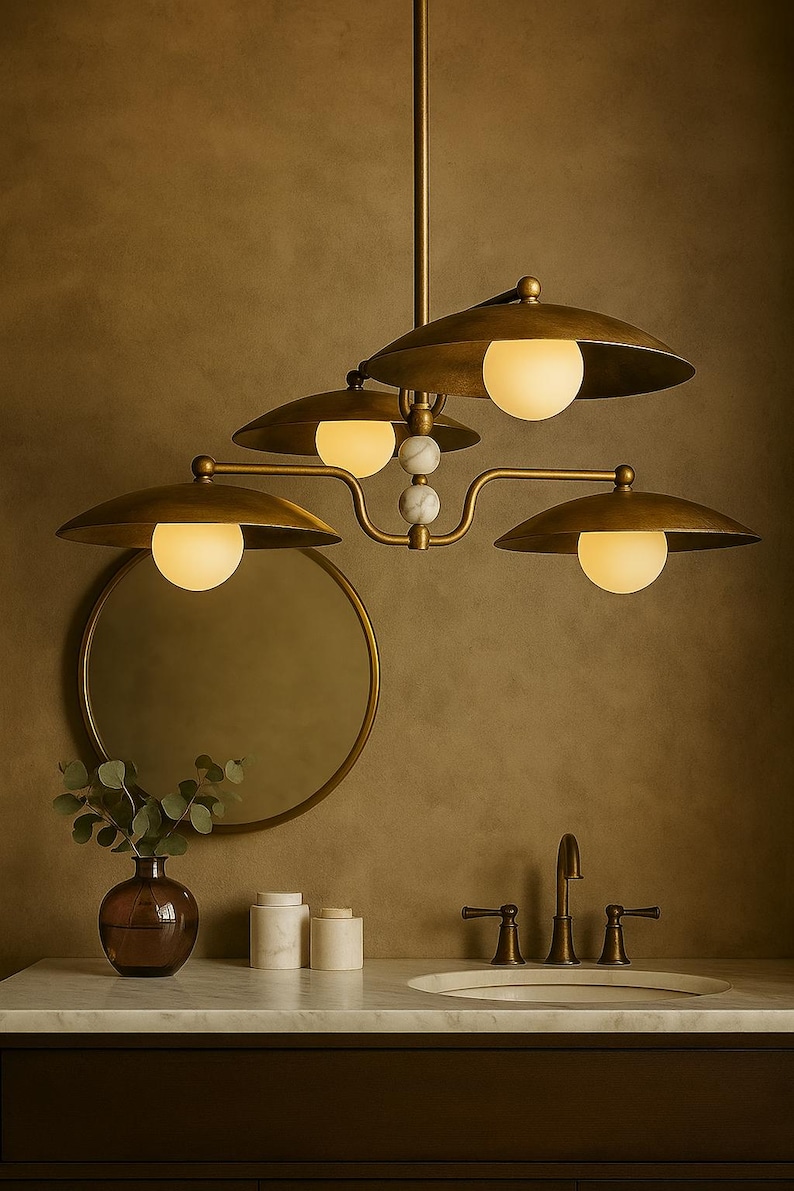 May include: A brass-toned chandelier with four lampshades and globe lights. The fixture has a central stem and curved arms. The lampshades are a flat, disc shape. The fixture is above a bathroom vanity with a mirror and sink.