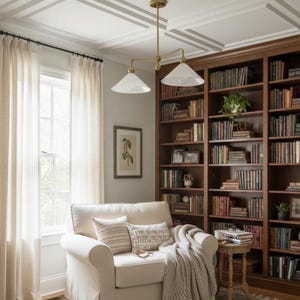May include: A comfortable reading area with a white armchair, cushions, and a throw. A large wooden bookcase filled with books is in the background. Sheer curtains frame a window, and a brass light fitting hangs from the ceiling.
