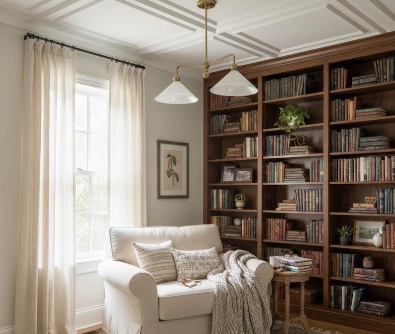 May include: A comfortable reading area with a white armchair, cushions, and a throw. A large wooden bookcase filled with books is in the background. Sheer curtains frame a window, and a brass light fitting hangs from the ceiling.