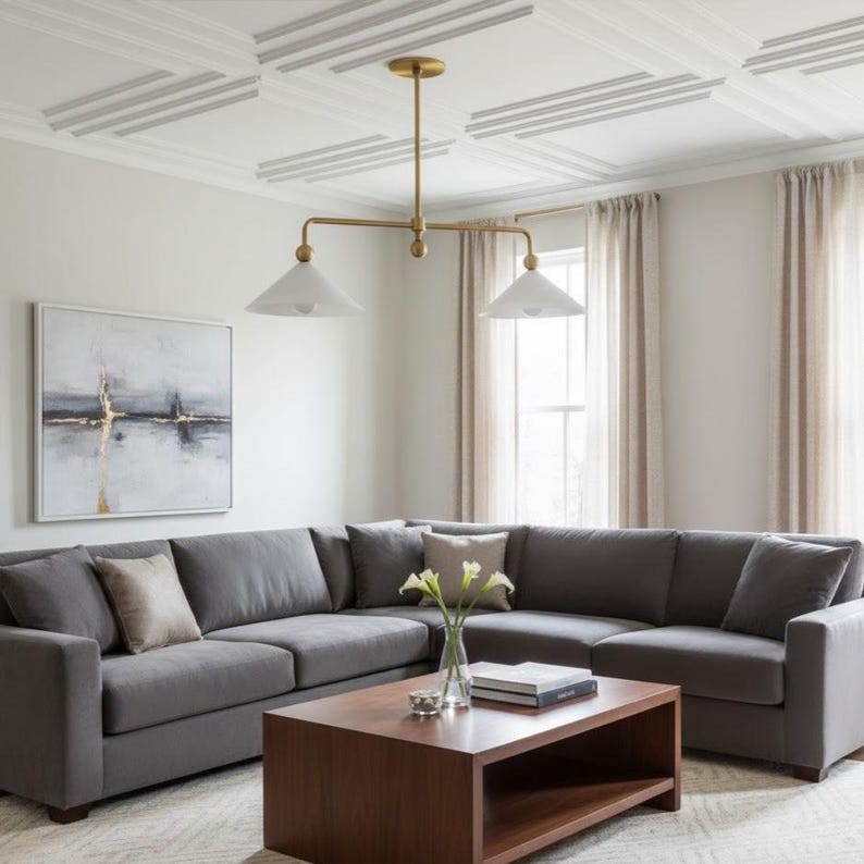 May include: A contemporary living room with a large grey sectional sofa, a dark wood coffee table, and a gold-toned chandelier with white shades. The room has neutral-coloured walls, curtains, and a patterned rug, creating a cosy atmosphere.