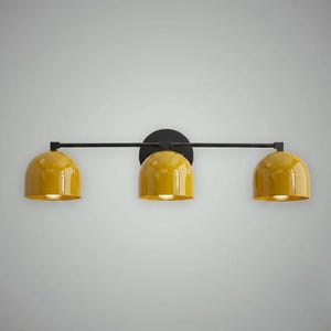 以下が含まれることがあります： 黒い金属フレームと円形の取り付けプレートを備えた、3灯の照明器具。この器具は、3つの黄色のドーム型のシェードを備えています。各シェードには電球が収められており、空間を照らします。デザインはモダンでミニマリストです。