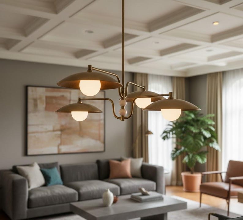 May include: A modern brass chandelier with four downward-facing, saucer-shaped shades, each housing a spherical light. The fixture hangs from a ceiling with a coffered design. The room features a gray sofa, artwork, and a large potted plant.