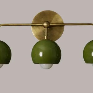 Op de afbeelding: Een wandlamp met drie lichtpunten, voorzien van een ronde messing achterplaat en een horizontale messing arm. Drie glanzende olijfgroene koepelvormige kappen zijn aan de arm bevestigd, elk met een kleine witte lamp die naar beneden schijnt.