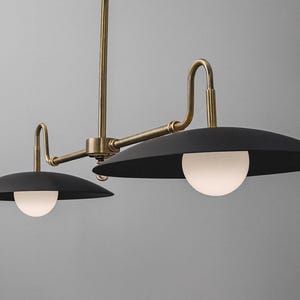May include: A modern, two-light pendant lamp with a brass finish and black shades. Each shade has a white, spherical light. The fixture is suspended from a central rod and has curved arms.