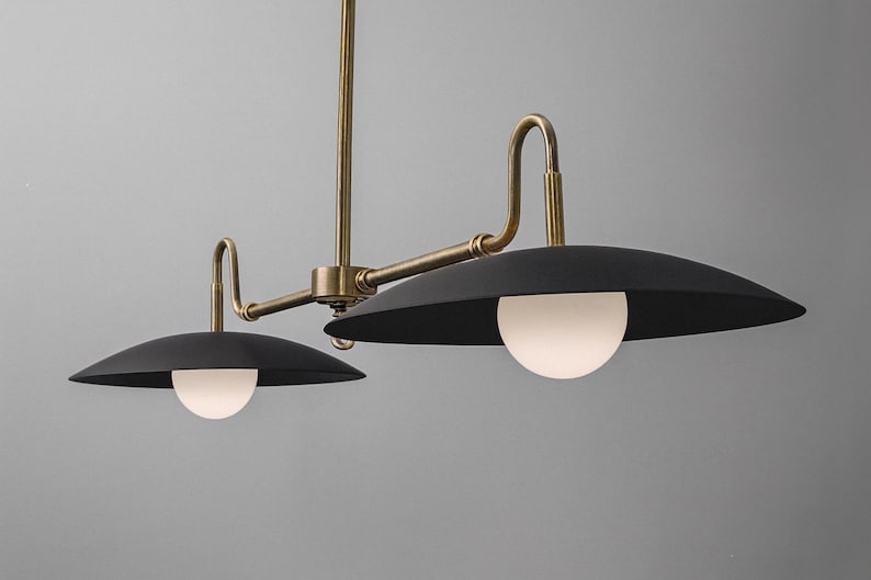 May include: A modern, two-light pendant lamp with a brass finish and black shades. Each shade has a white, spherical light. The fixture is suspended from a central rod and has curved arms.