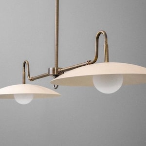 May include: A two-light pendant light fixture with a vintage brass finish. The fixture features two off-white, saucer-shaped shades with a white globe light in the center of each. The fixture is suspended from a central rod.
