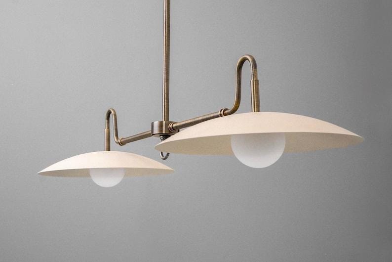 May include: A two-light pendant light fixture with a vintage brass finish. The fixture features two off-white, saucer-shaped shades with a white globe light in the center of each. The fixture is suspended from a central rod.