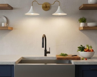 May include: A modern kitchen sink with a black faucet, white countertops, and navy blue cabinets. Above the sink, a gold-toned light fixture with two white cone-shaped shades illuminates the space. Wooden shelves and a bowl of produce add warmth.