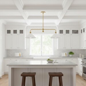 May include: A modern kitchen with white cabinets, countertops, and ceiling beams. A gold-toned light fixture hangs above a kitchen island with two brown wooden stools. A stainless steel oven and range are visible.