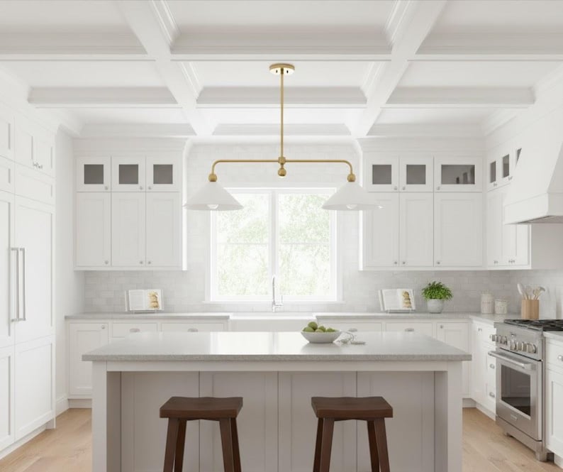 May include: A modern kitchen with white cabinets, countertops, and ceiling beams. A gold-toned light fixture hangs above a kitchen island with two brown wooden stools. A stainless steel oven and range are visible.