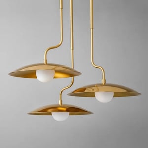 May include: Three-light pendant fixture with a gold finish. Each light features a curved arm, a circular shade, and a white globe-shaped bulb. The fixture is suspended from the ceiling.