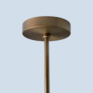 May include: A close-up of a bronze-colored ceiling light fixture. The fixture has a circular canopy and a slender rod extending downwards. The light fixture is against a light blue background.