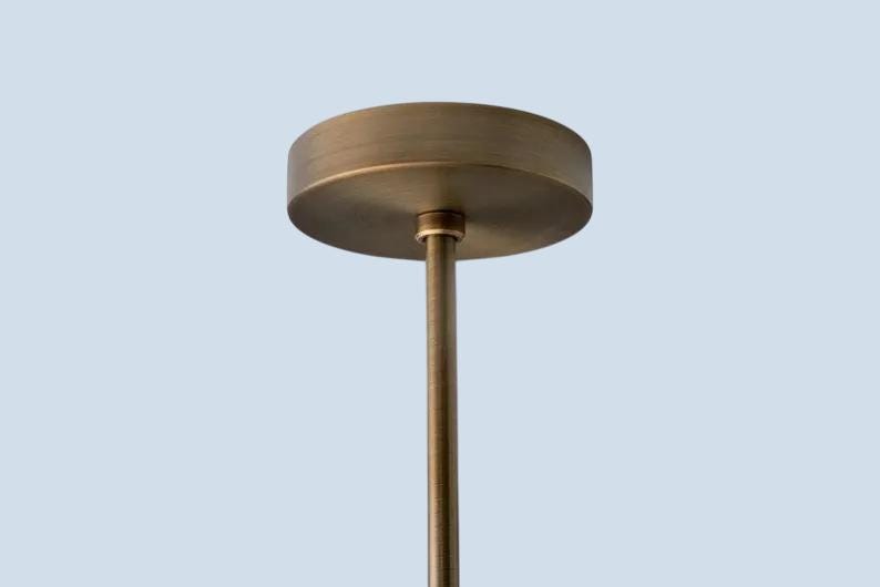 May include: A close-up of a bronze-colored ceiling light fixture. The fixture has a circular canopy and a slender rod extending downwards. The light fixture is against a light blue background.