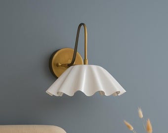 May include: A wall-mounted light fixture with a brass finish and a white, ruffled shade. The fixture has a circular base and a curved arm supporting the shade. The shade has a wavy, scalloped edge. The light is mounted on a blue wall.