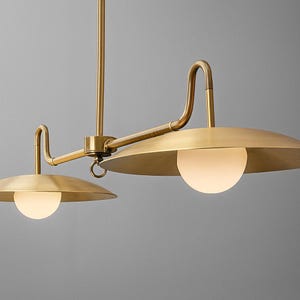 May include: A brass-colored, two-light pendant lamp with a minimalist design. The lamp features two circular shades with a flat, disc-like shape, each housing a spherical, frosted glass light. The fixture has curved arms and a central support.