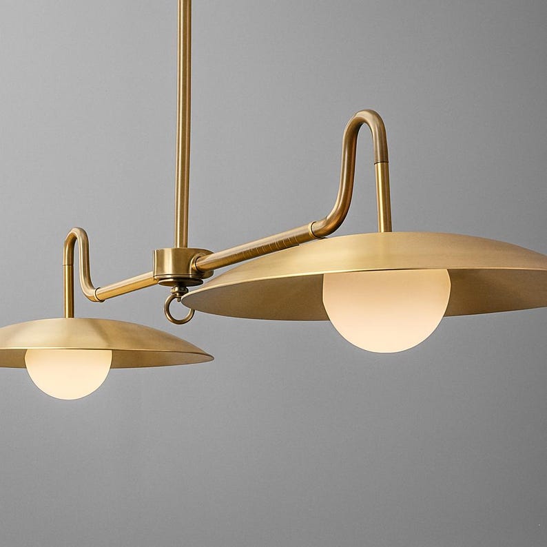 May include: A brass-colored, two-light pendant lamp with a minimalist design. The lamp features two circular shades with a flat, disc-like shape, each housing a spherical, frosted glass light. The fixture has curved arms and a central support.