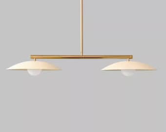 May include: A modern, two-light pendant lamp with a gold-toned metal frame. The lamp features two off-white, saucer-shaped shades with a spherical light bulb in each. The design is minimalist and elegant, suitable for various interior styles.
