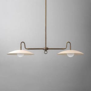 May include: A brass-toned, two-light pendant light fixture. The fixture has a central vertical rod with a horizontal bar extending to each side. Each side has a curved arm holding a cream-colored, saucer-shaped shade with a white globe bulb.