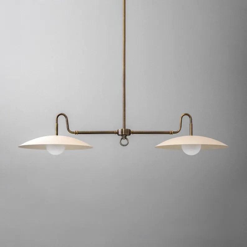 May include: A brass-toned, two-light pendant light fixture. The fixture has a central vertical rod with a horizontal bar extending to each side. Each side has a curved arm holding a cream-colored, saucer-shaped shade with a white globe bulb.