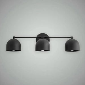 以下が含まれることがあります： 黒色の3灯式洗面化粧台用照明器具。各電球を備えた3つのドーム型のシェードがあります。シェードは水平バーに取り付けられ、円形のベースに接続されています。壁への取り付け用に設計されています。