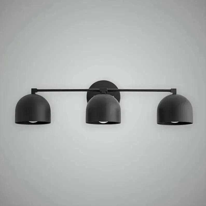 以下が含まれることがあります： 黒色の3灯式洗面化粧台用照明器具。各電球を備えた3つのドーム型のシェードがあります。シェードは水平バーに取り付けられ、円形のベースに接続されています。壁への取り付け用に設計されています。