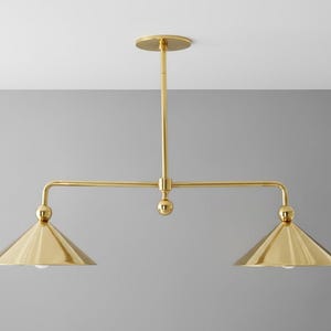 May include: A gold-coloured, two-light pendant light fitting. The fitting has a central vertical rod attached to a circular ceiling mount. Two horizontal arms extend from the central rod, each supporting a conical shade with a bulb. The fitting is made of metal.