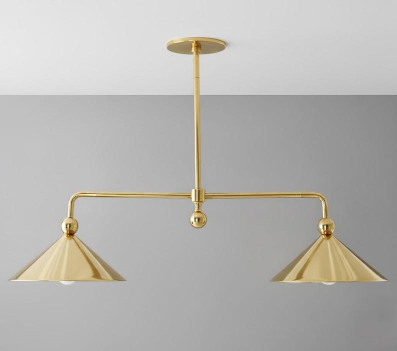 May include: A gold-coloured, two-light pendant light fitting. The fitting has a central vertical rod attached to a circular ceiling mount. Two horizontal arms extend from the central rod, each supporting a conical shade with a bulb. The fitting is made of metal.