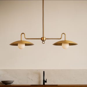 May include: A brass-toned, two-light pendant light fixture. The fixture has a central vertical rod with a horizontal bar extending to two curved arms, each supporting a downward-facing, saucer-shaped shade and a spherical light bulb. The design is minimalist and modern.