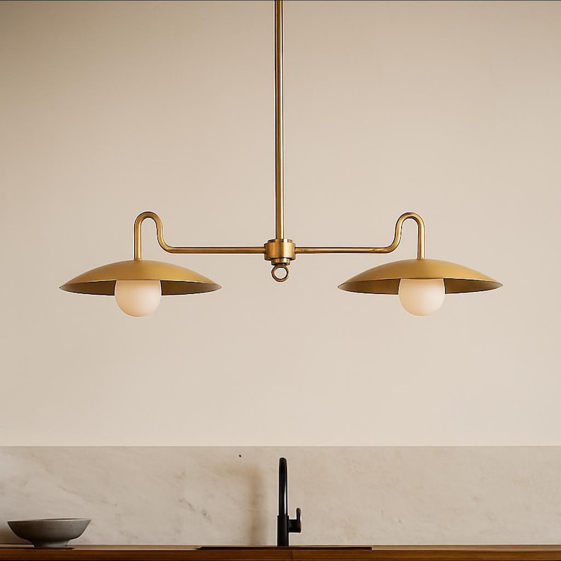 May include: A brass-toned, two-light pendant light fixture. The fixture has a central vertical rod with a horizontal bar extending to two curved arms, each supporting a downward-facing, saucer-shaped shade and a spherical light bulb. The design is minimalist and modern.