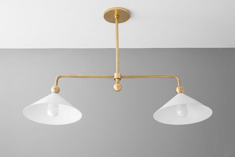 May include: A contemporary pendant light with two lights. The fixture has a gold-coloured metal frame and two white, cone-shaped shades. It is designed to be mounted on a ceiling and is suitable for various interior styles.