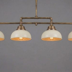Könnte beinhalten: Eine Pendelleuchte mit vier Lichtern im Vintage-Industriedesign. Die Leuchte verfügt über vier cremefarbene, kuppelförmige Schirme mit freiliegenden Glühbirnen. Der Metallrahmen hat eine Bronzefinish und die Lampe hängt an einer zentralen Stange.