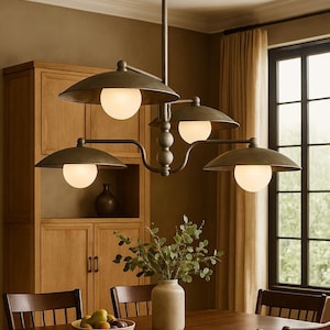May include: A bronze-finished chandelier with four lights, each encased in a white globe, hangs above a wooden dining table. The fixture has a classic, industrial design. The dining room features a wooden cabinet, chairs, and a vase with greenery.